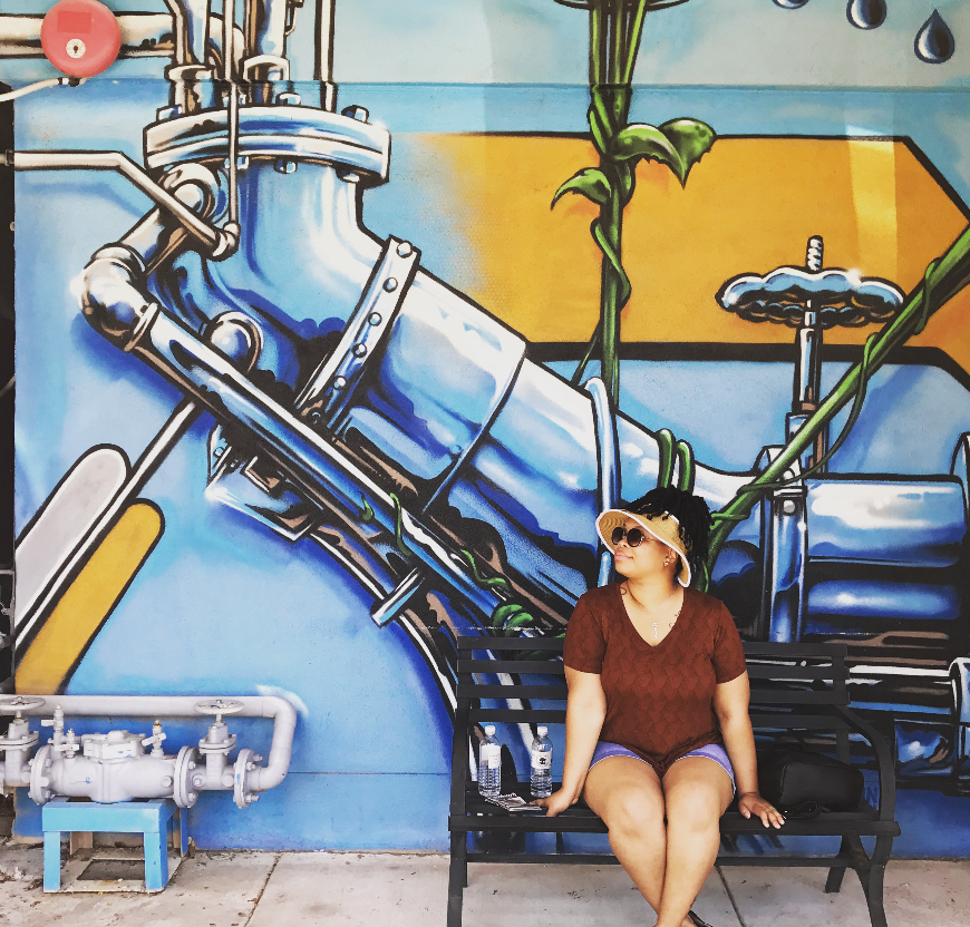 Woman sitting on a bench in front of a colorful graffiti mural of industrial pipes and valves.