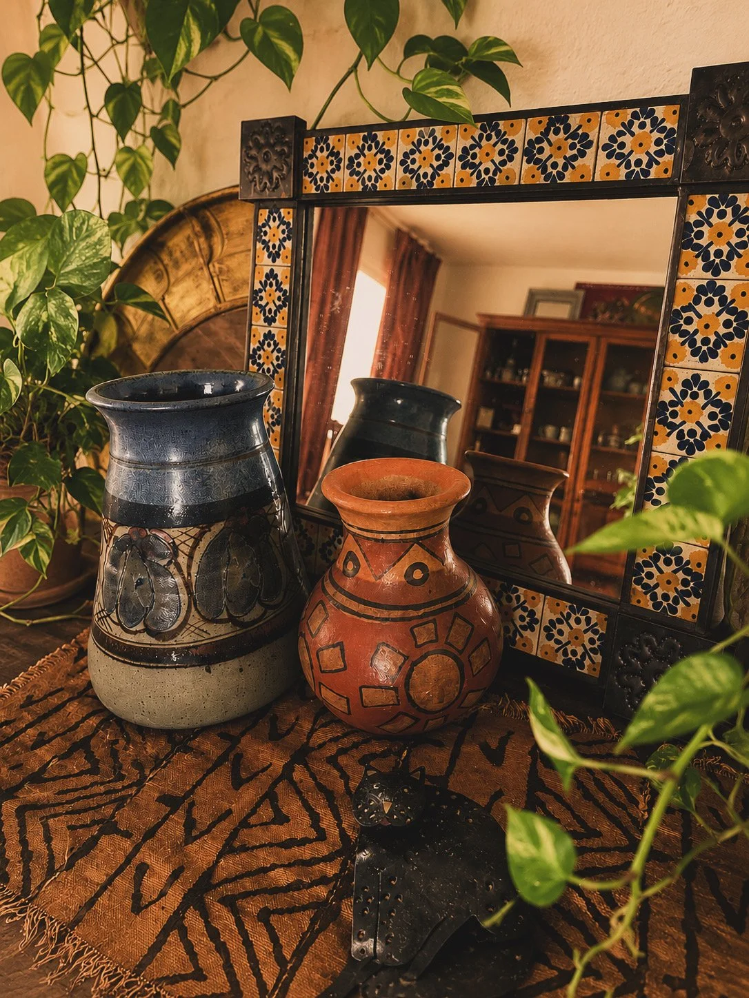 Decorative pottery vases and a mirror with tile border on a table with a cloth, surrounded by green plants in a cozy home interior.