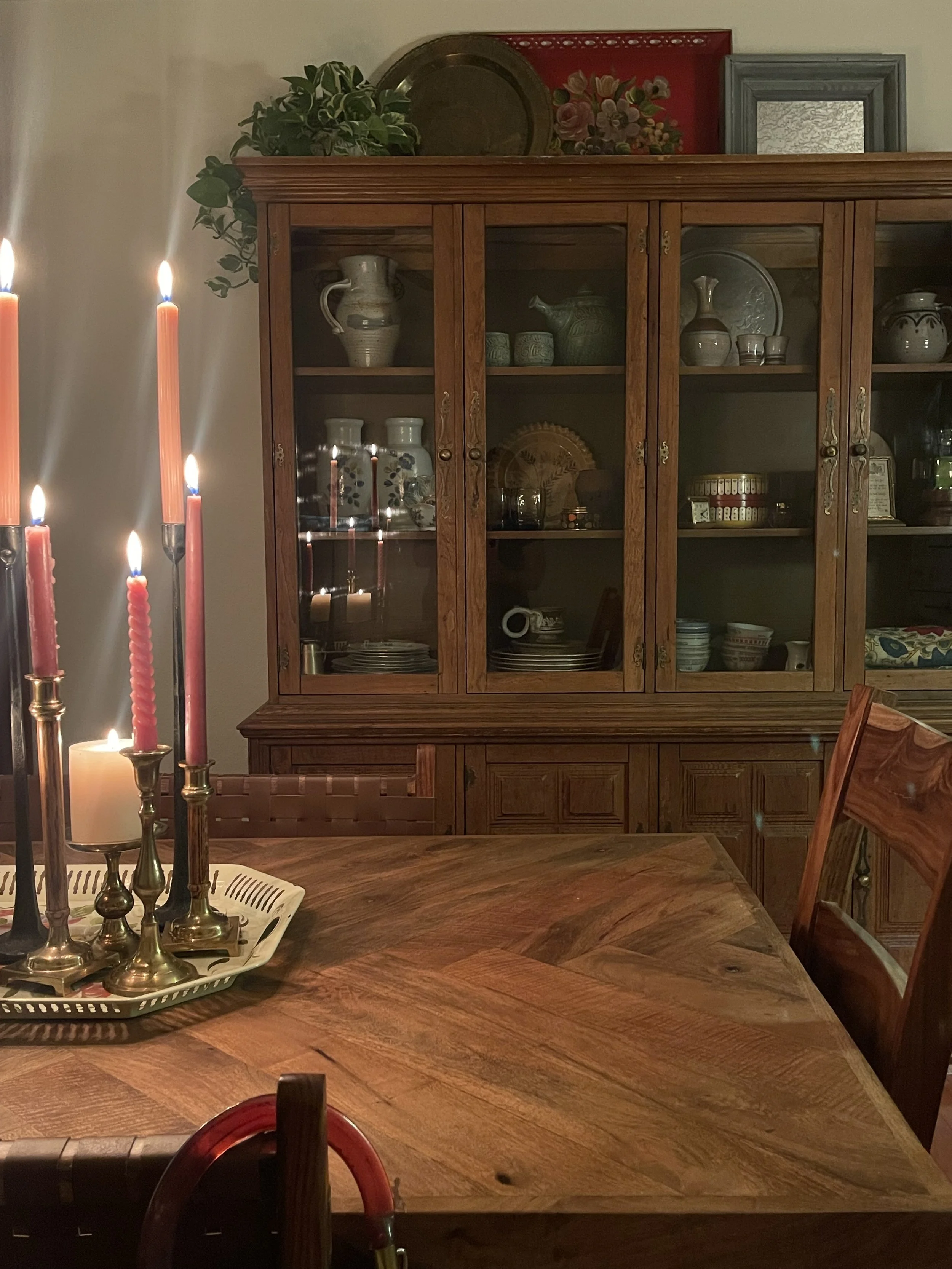 Dining room scene with a wooden table, red candles with flames, and a wooden china cabinet filled with dishes, pitchers, and decorative items. The cabinet is topped with artwork and a potted plant.