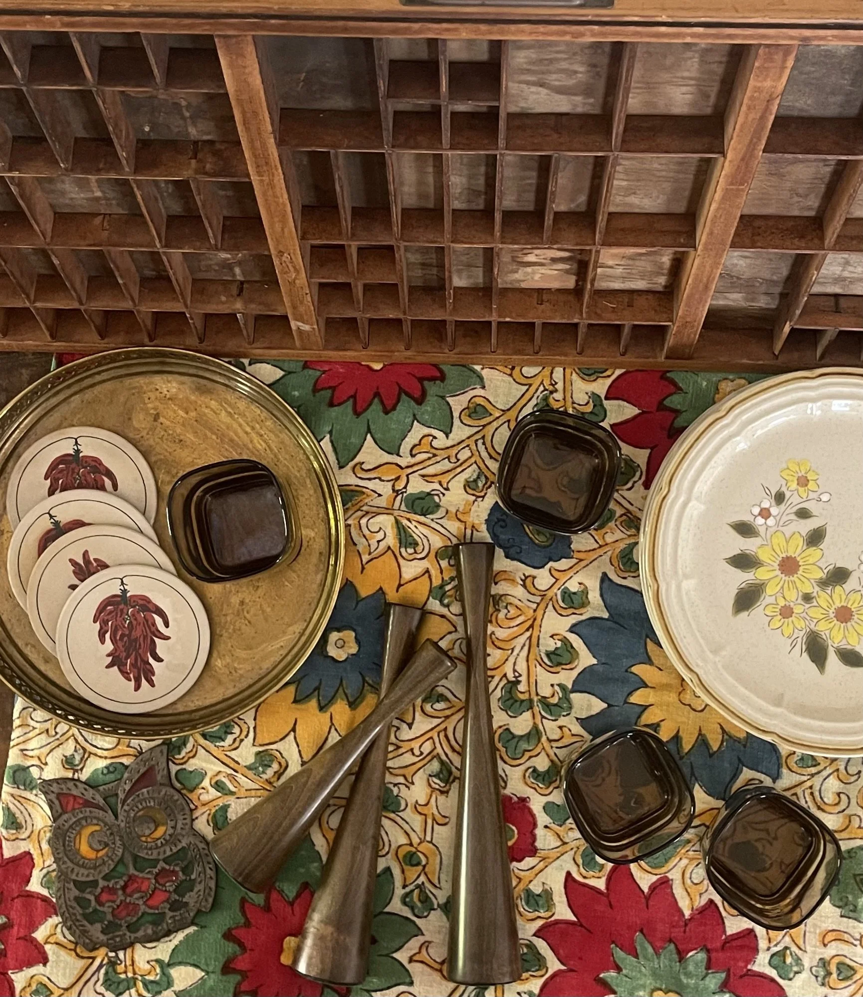 View of a wooden cupcake or muffin holder with several compartments, placed on a colorful floral tablecloth. There are also some small black containers and a decorative plate with a yellow flower design.
