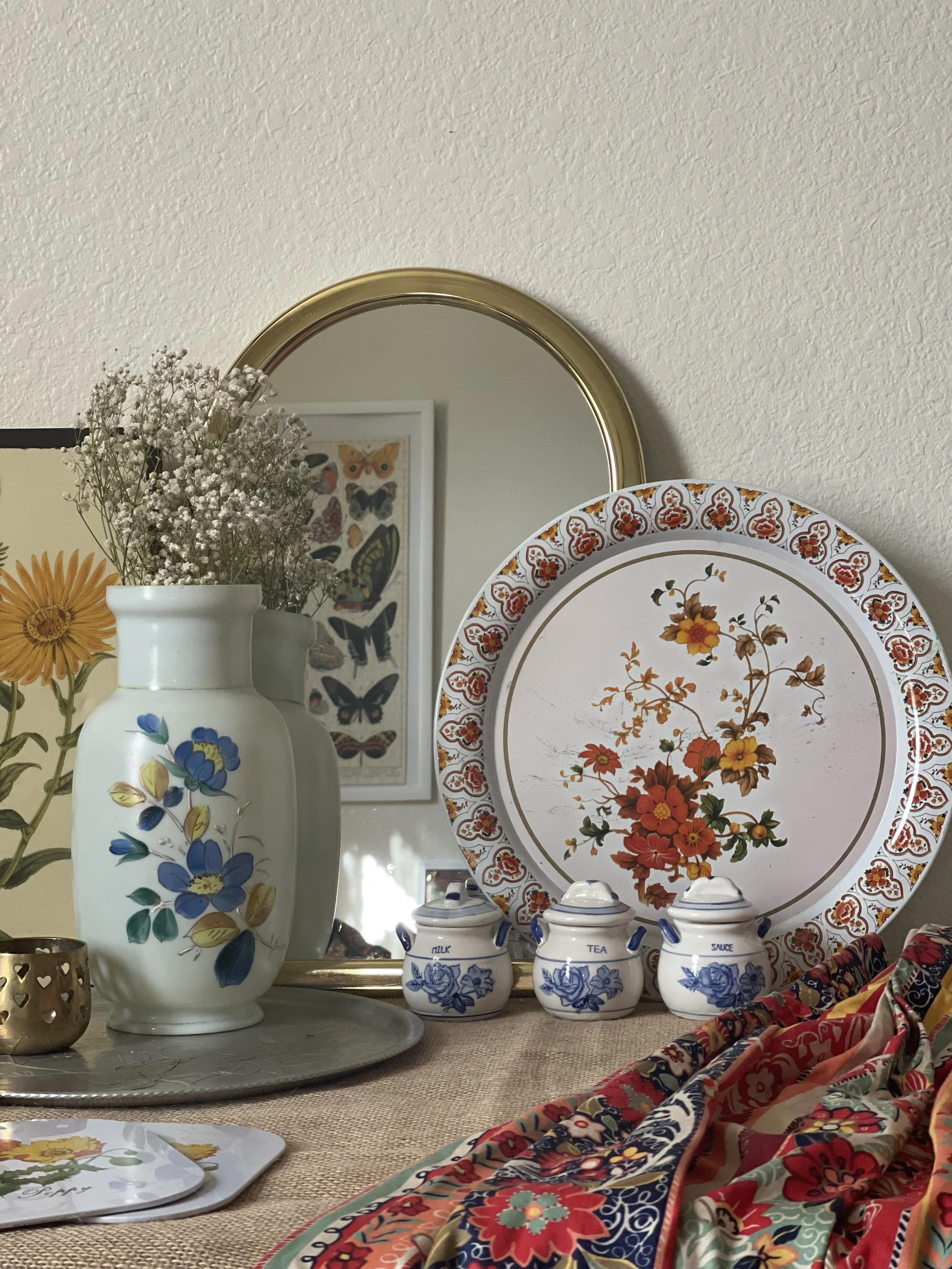 A decorative tabletop arrangement featuring a tall white ceramic vase with painted blue and yellow flowers, filled with small white flowers. Behind it, a round mirror with a gold rim reflects a framed butterfly illustration on the wall. To the right, a large floral-patterned tray leans against the wall. In front, there are three small ceramic containers labeled 'Milk,' 'Tea,' and 'Sauce,' and a colorful, floral cloth draped over the edge of the table.