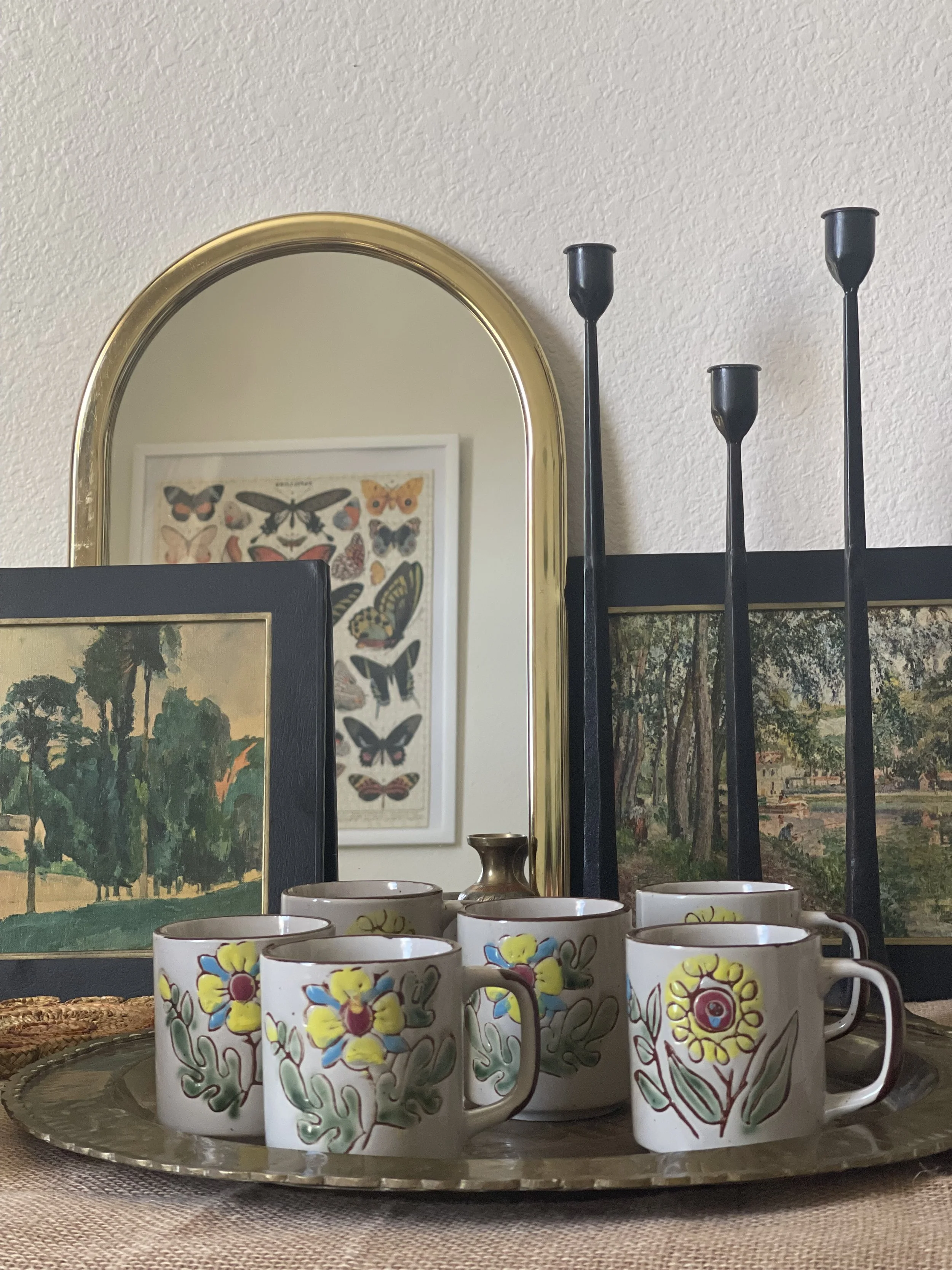 A silver tray with floral patterned mugs, a small brass container, and a group of painted mugs on a woven placemat, with framed butterfly illustrations and landscape paintings in the background.