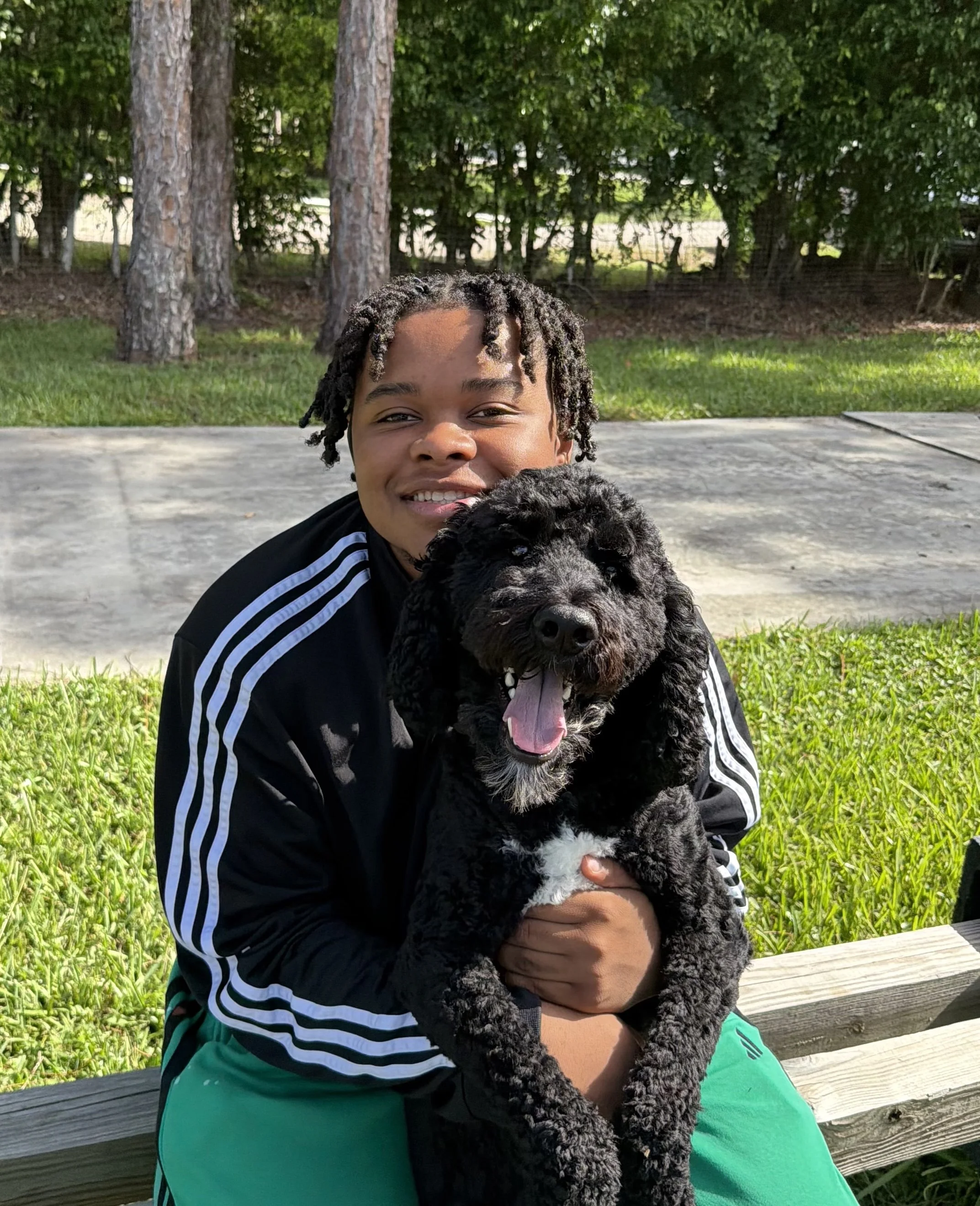 A person with styled dreadlocks smiling and praising a fluffy black puppy while sitting on a wooden bench outdoors in a park with green grass, trees, and bushes in the background.