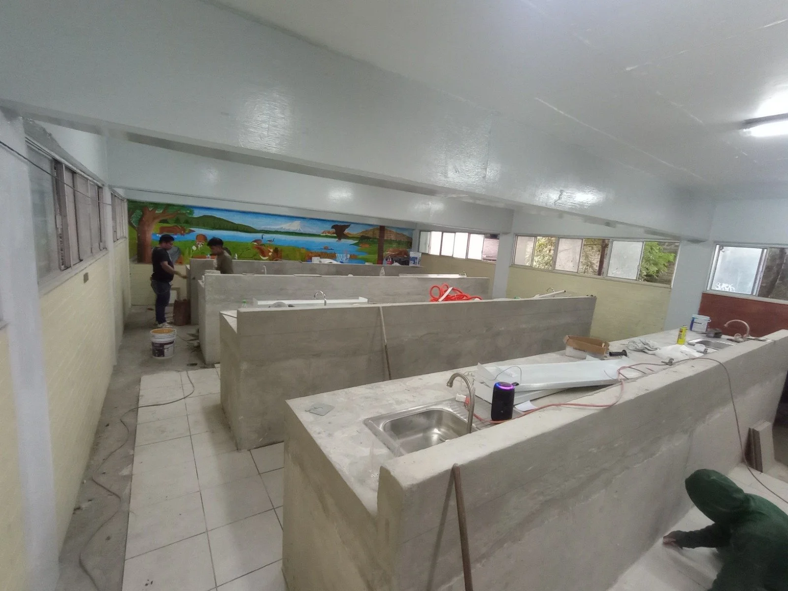 Área en construcción con cabinas divididas, fregaderos, y trabajadores instalando accesorios. Decoración en la pared con mural de paisaje natural.