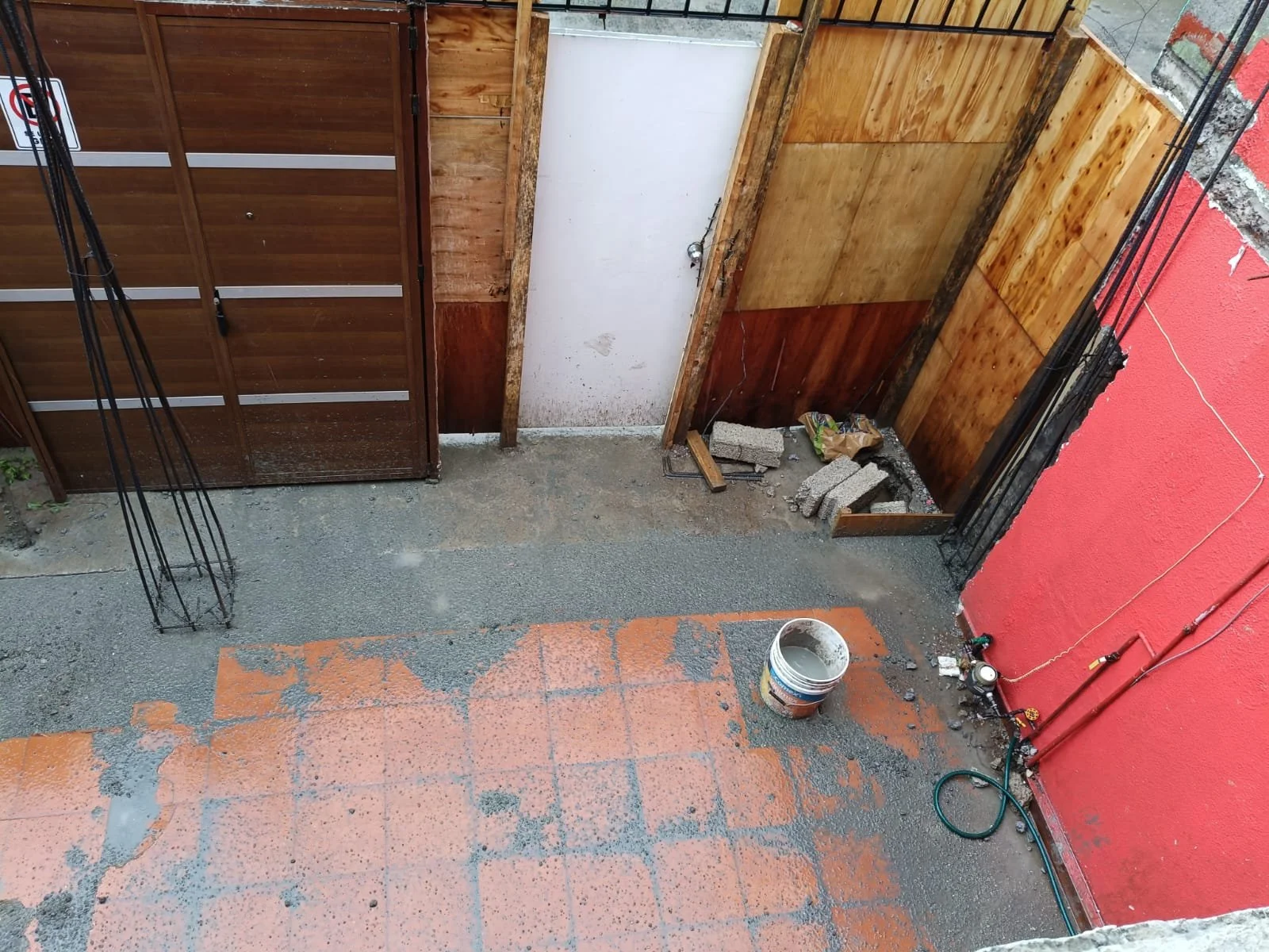 Área en construcción con pared de bloque y madera, puerta blanca, suelo de concreto con manchas de pintura, cubeta de pintura y materiales de construcción en el piso, detrás de una reja de barrotes negro.