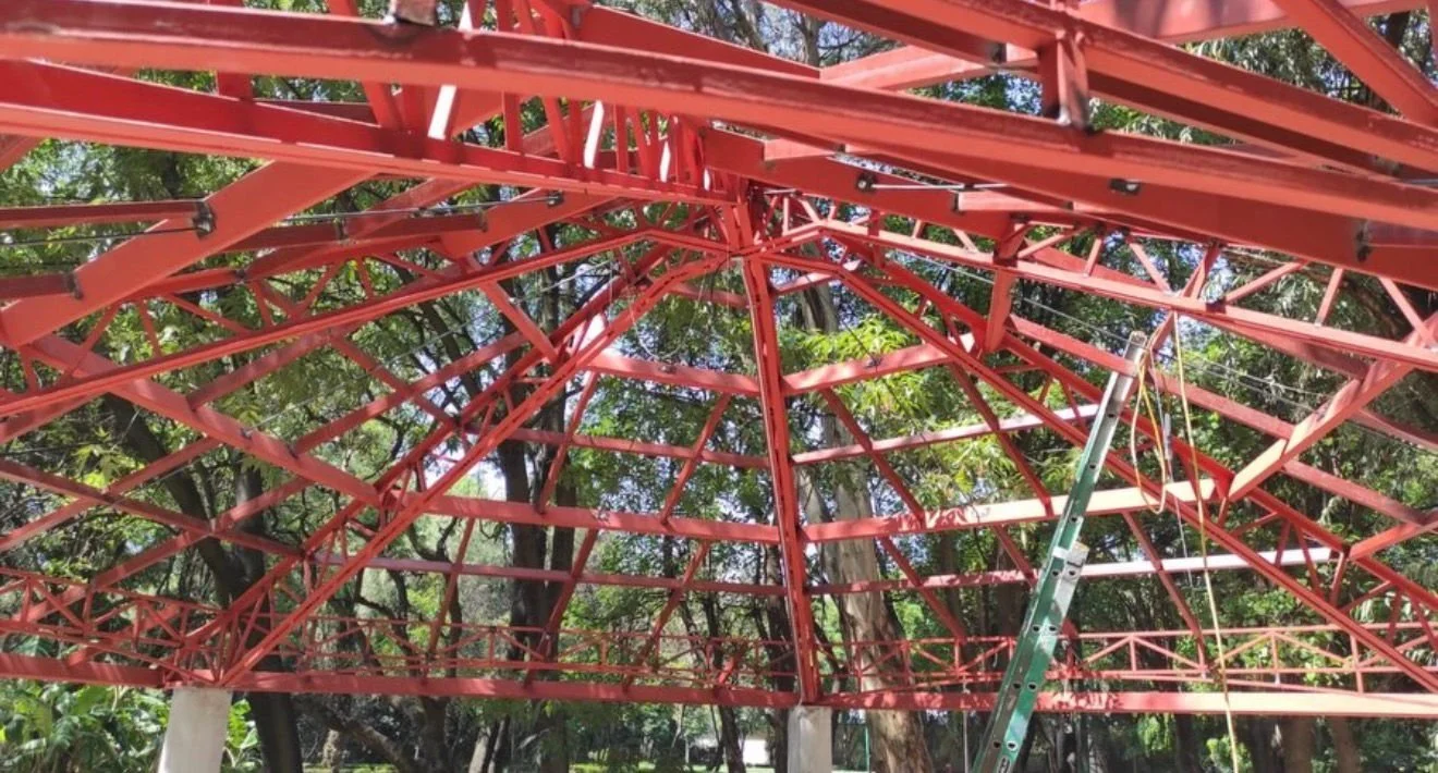 Estructura metálica roja en construcción, con un andamio y árboles de fondo.