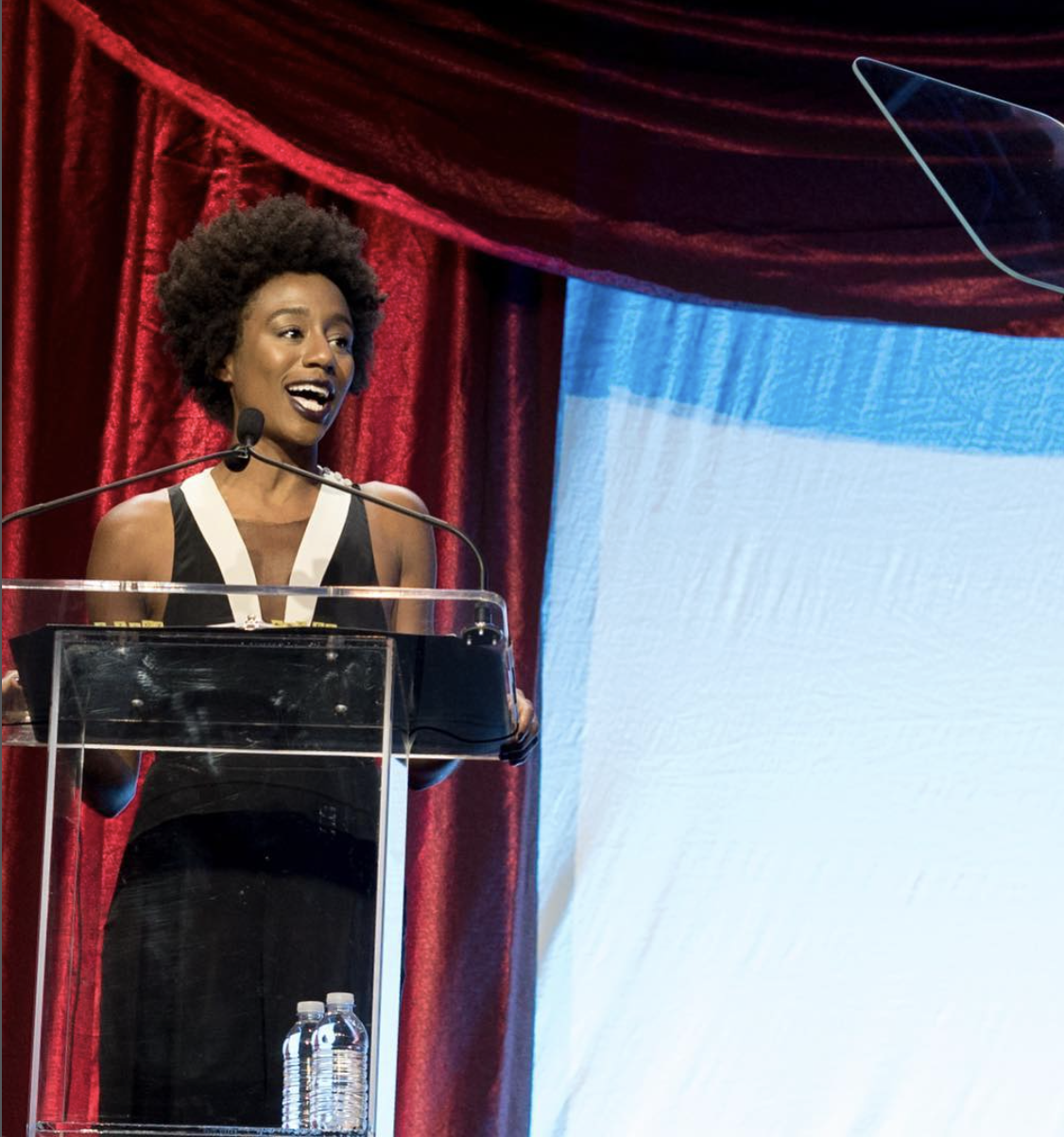 A woman speaking at a podium on a stage with red curtains and a blue and white backdrop.