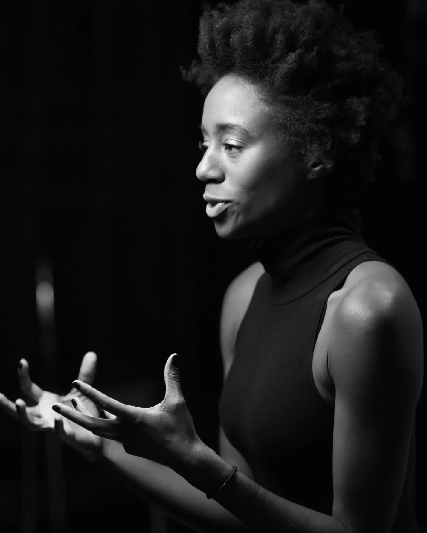 A black and white photo of a woman speaking or explaining, with her hands gesturing in front of her. She has a serious expression, short curly hair, and is wearing a sleeveless top and a choker.