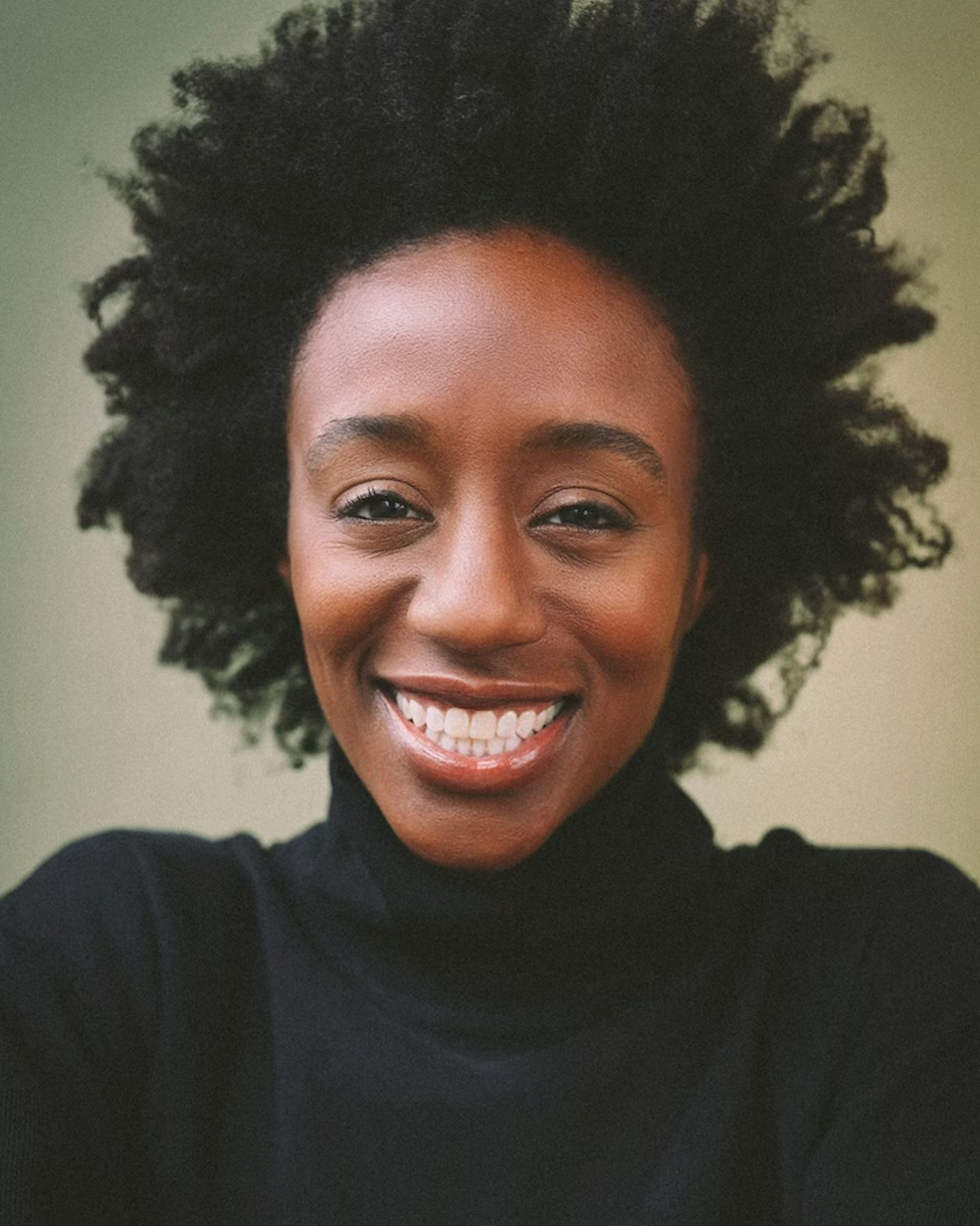 Close-up portrait of a smiling Black woman with natural curly hair, wearing a black turtleneck against a plain background.