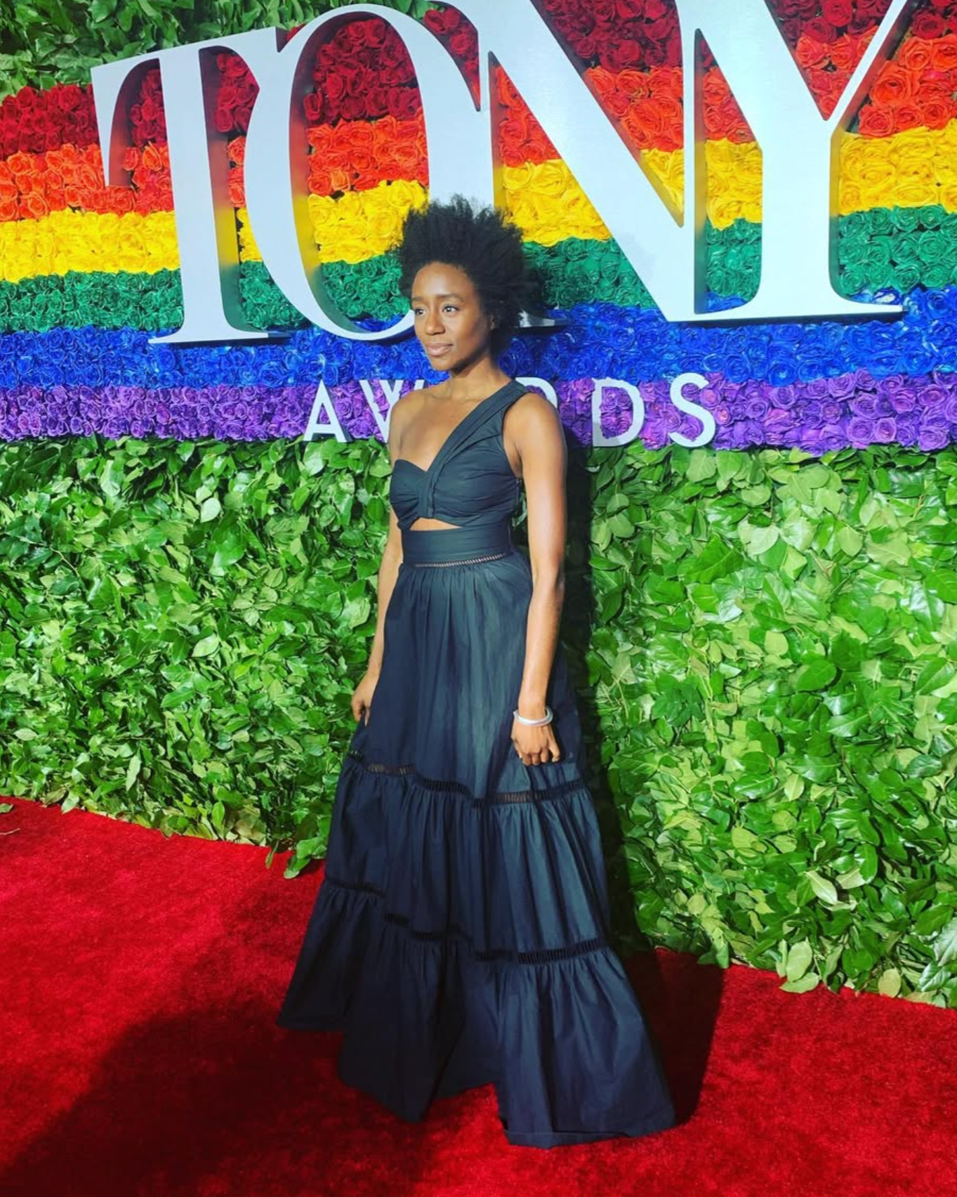 A woman in a dark blue, off-shoulder gown stands on a red carpet in front of a colorful floral backdrop with the words 'ICON AWARDS' displayed. The backdrop features green leaves and a rainbow of roses in red, orange, yellow, green, blue, purple, and