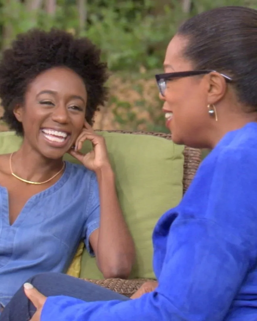 Two women are sitting outdoors, smiling and talking. The woman on the left has short, curly hair, is wearing a blue top, and a gold necklace. The woman on the right has glasses, pulled-back hair, wearing a blue top, and earrings.
