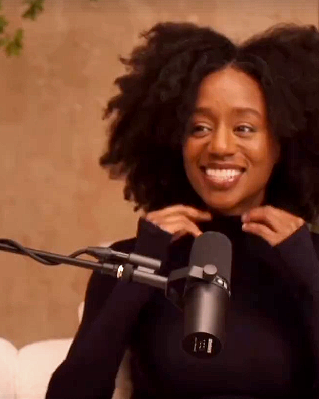 A woman with curly black hair smiling while sitting in front of a microphone.
