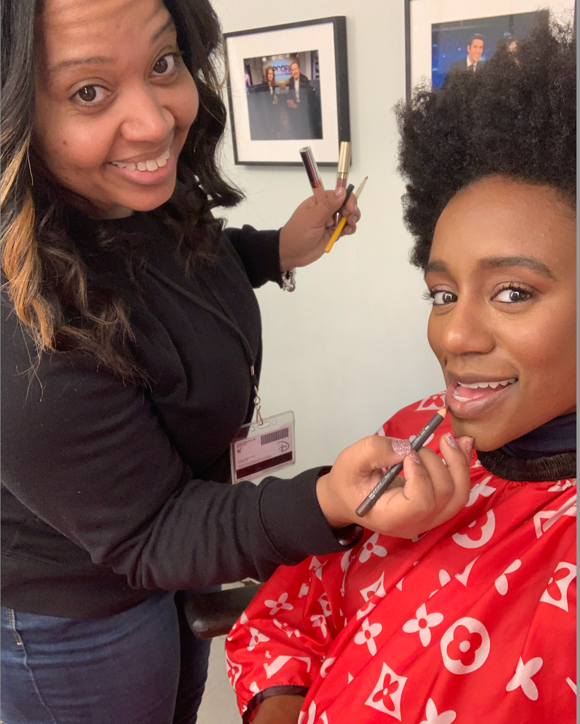A makeup artist applying lipstick to a woman with an afro hairstyle, who is wearing a red jacket with white floral patterns. The makeup artist is smiling and holding makeup brushes, while the woman is smiling at the camera.