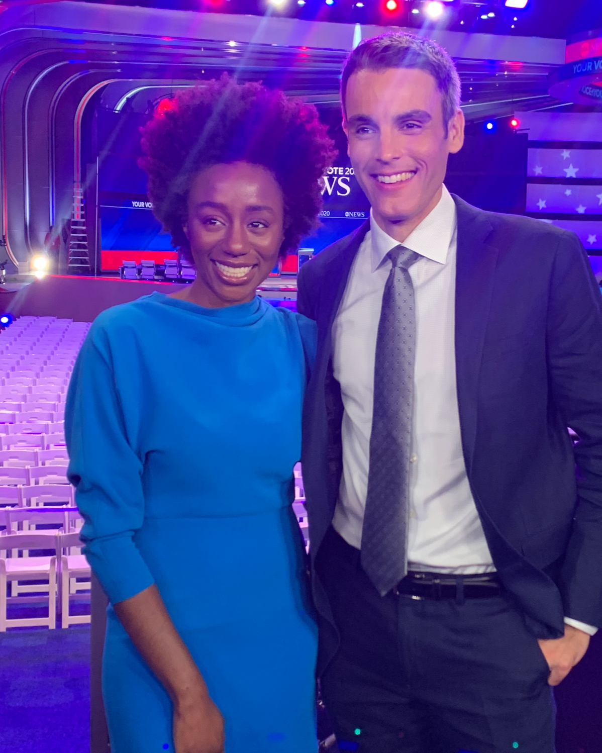 A woman with curly hairstyle in a blue dress standing next to a man wearing a suit and tie, both smiling, in a large, colorful, well-lit auditorium or stage with chairs and a backdrop displaying news graphics.