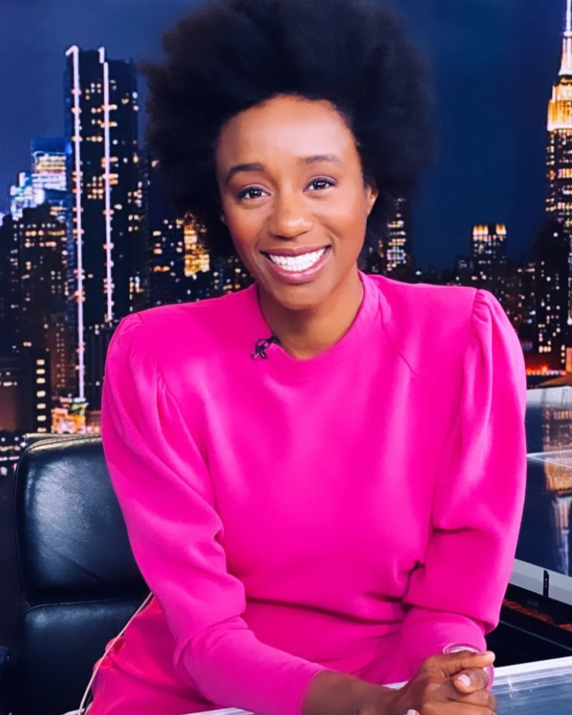 A woman with dark curly hair smiling, wearing a bright pink top, sitting in front of a city skyline backdrop at night.