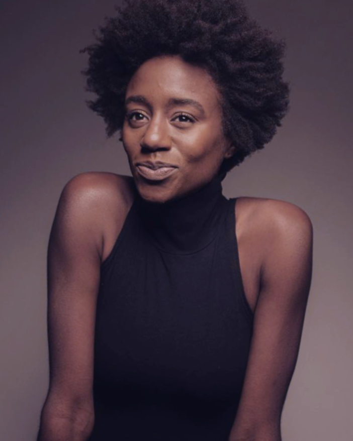 Portrait of a woman with natural afro hair, wearing a sleeveless black turtleneck, smiling softly against a neutral background.