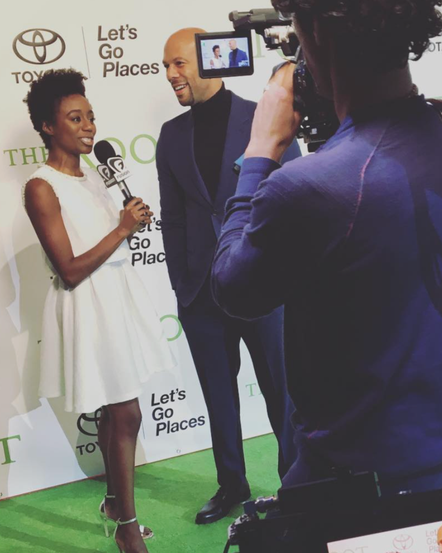 A woman being interviewed on a red carpet with a microphone, standing next to a man in a blue suit. They are both smiling, and a cameraman is recording them. The backdrop features the Toyota logo and the words 'Let's Go Places'.