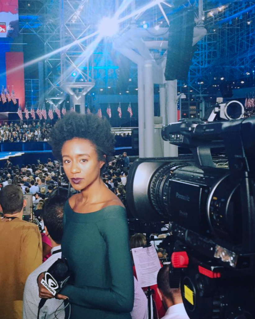 A female reporter holding a microphone, standing in front of a large crowd at a political event with American flags, inside a modern venue with blue lighting and scaffolding.