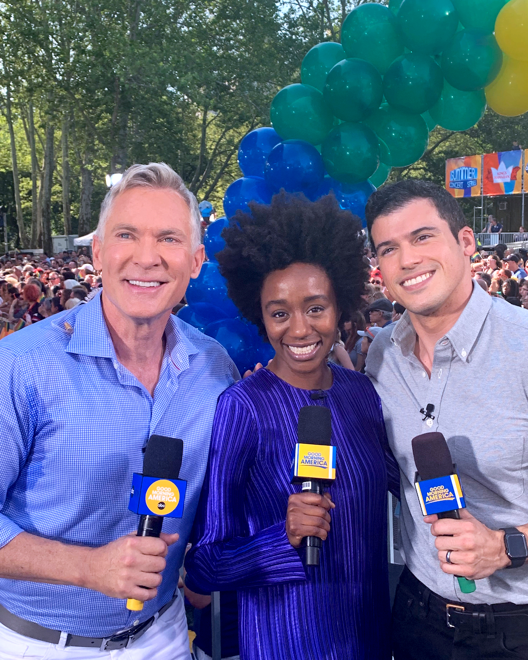 Three news anchors smiling and holding microphones with 'Good Morning America' logos, standing outdoors at a large crowd event with blue and green balloons in the background.