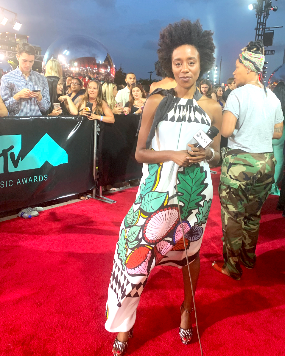 Woman with natural afro hairstyle holding a microphone and standing on a red carpet at the MTV Music Awards, wearing a colorful, patterned dress with floral and geometric motifs, surrounded by a crowd of people and event lighting.
