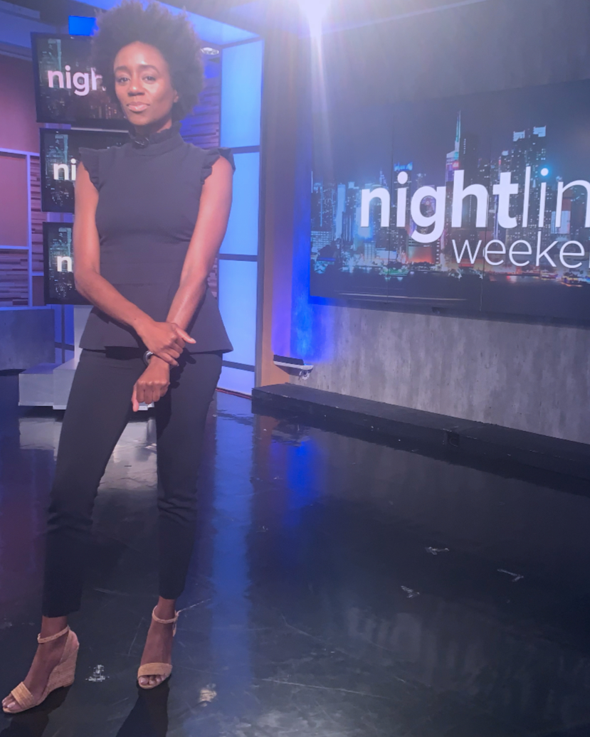 A woman standing on a TV studio set with a background screen displaying the words "nightline weekend" and a cityscape, wearing a sleeveless black outfit and nude heels.