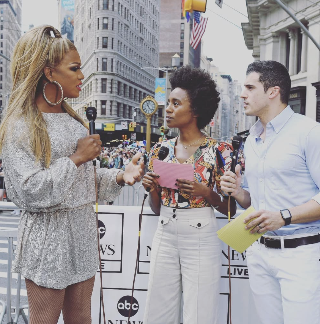 Three news reporters interviewing each other on a city street during a public event, with a crowd and tall buildings in the background.
