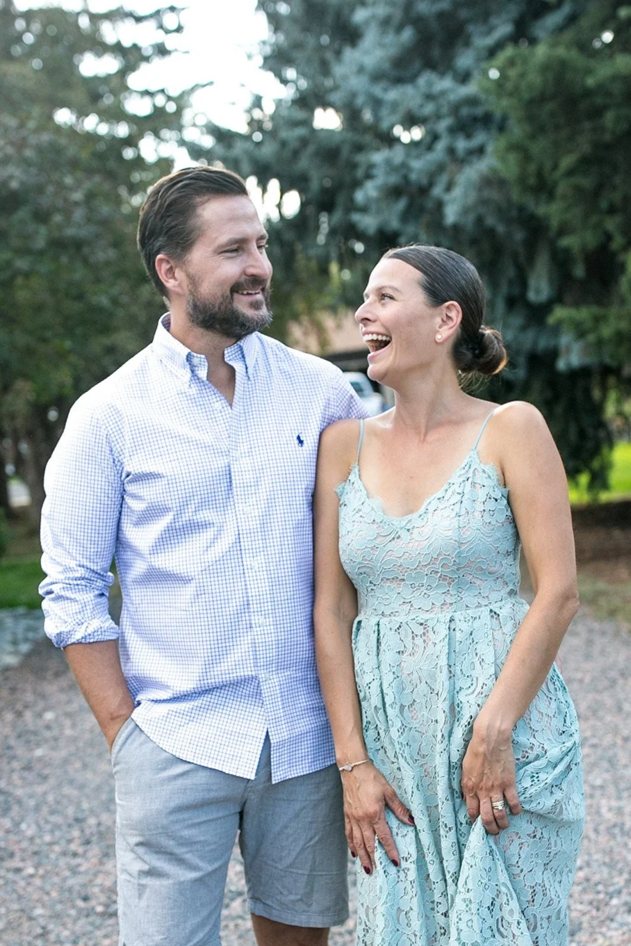A man and woman standing outdoors, smiling and looking at each other, with trees in the background.