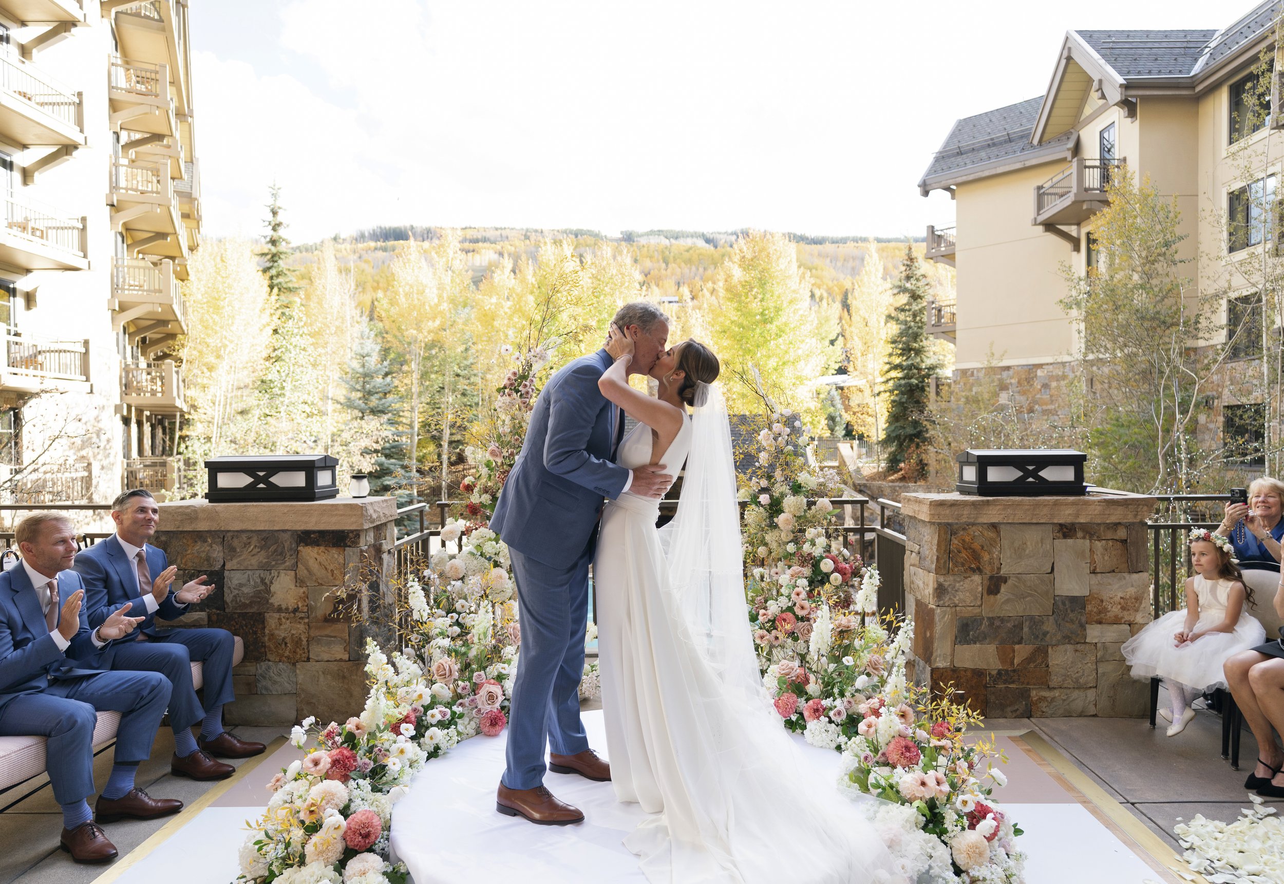 A couple kisses during a wedding ceremony outdoors, surrounded by guests seated on either side, with a backdrop of trees and buildings, decorated with numerous pink, white, and peach flowers.