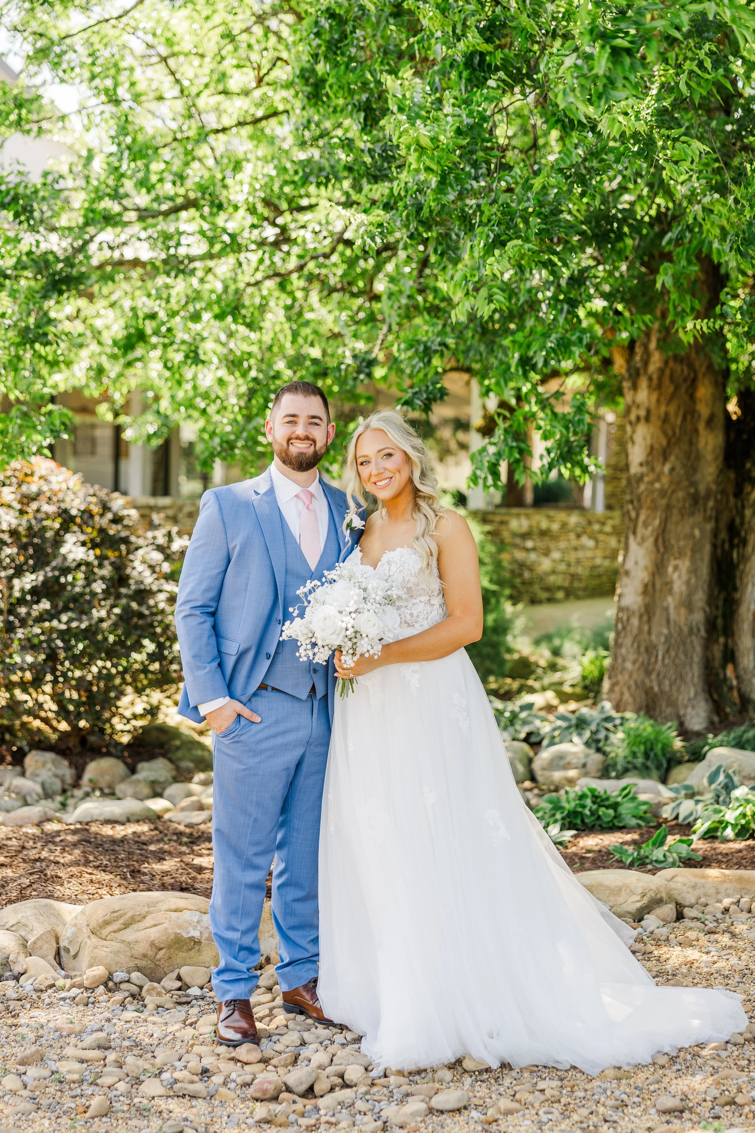 A newlywed couple standing outdoors on a rocky path in a garden, smiling and posing for a photo under a large leafy tree. The groom is wearing a light blue suit with a pink tie, and the bride is in a white wedding gown holding a bouquet of white flow