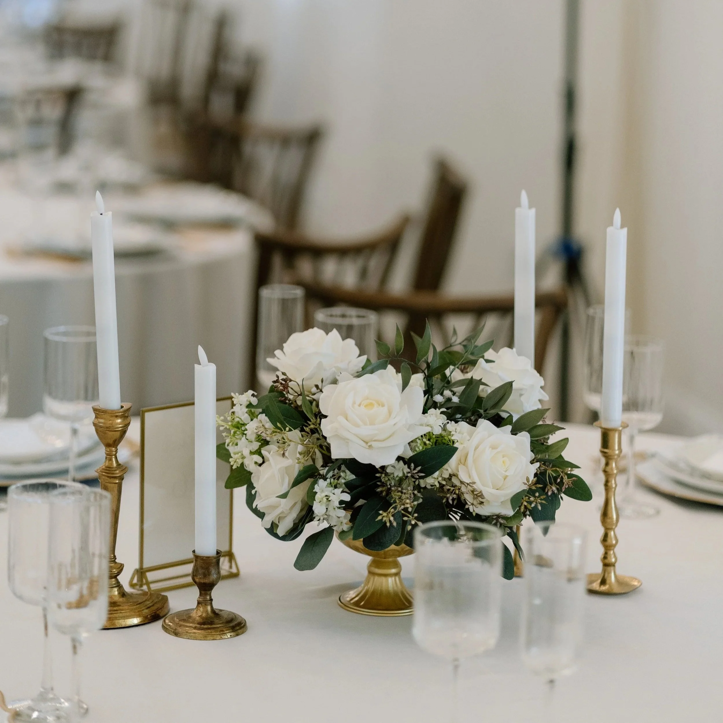 A table decorated with white roses, greenery, and white candles in gold holders, with a blurred background of chairs and part of a dining area.