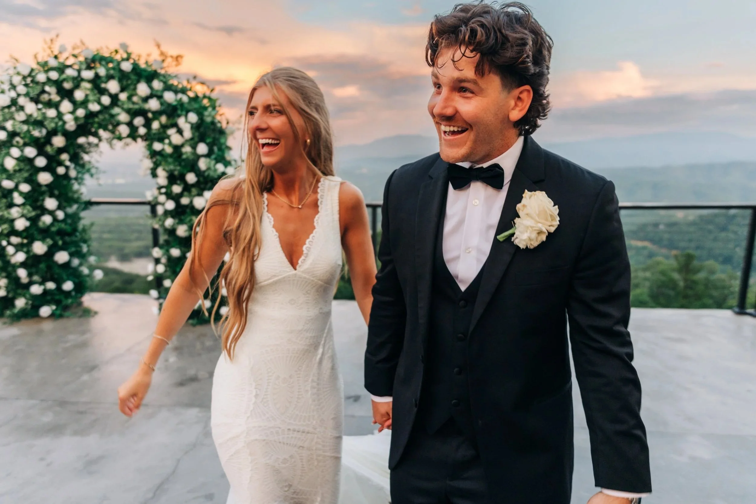 A smiling couple dressed in wedding attire, holding hands, walking outdoors at sunset with a scenic view and floral arch in the background.