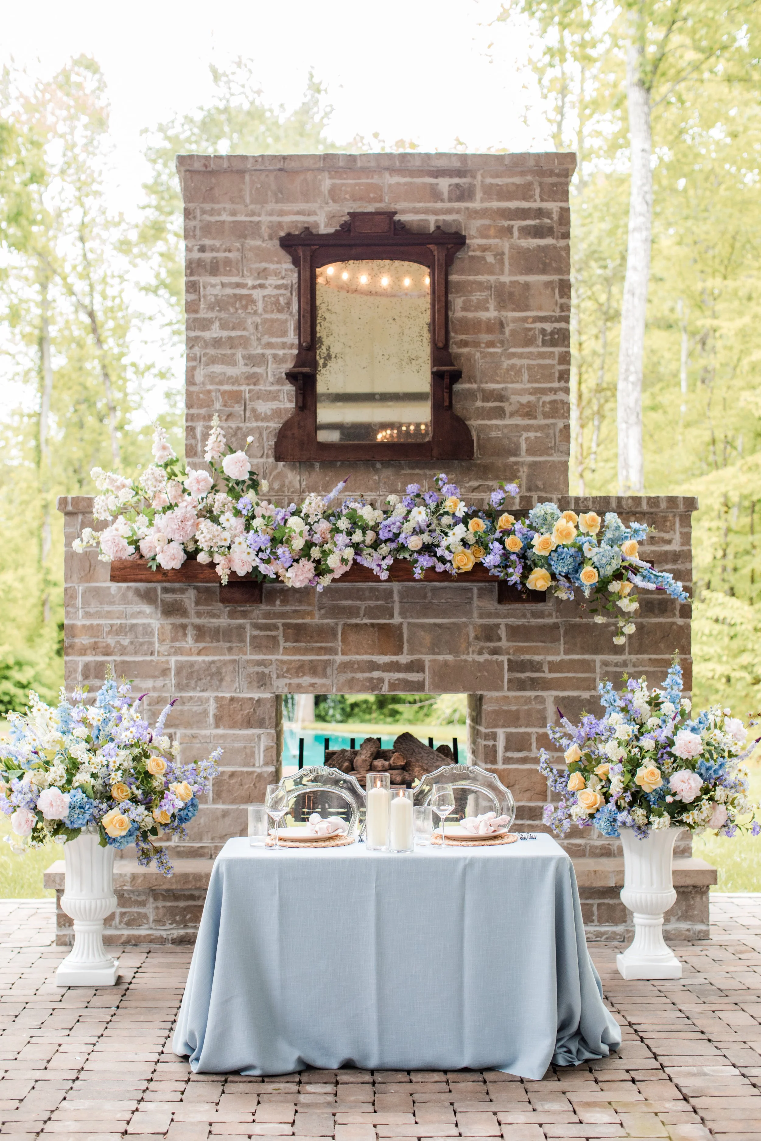 A decorated outdoor table set for a special occasion, with flowers, candles, and place settings in front of a brick fireplace with a mirror and floral arrangements.