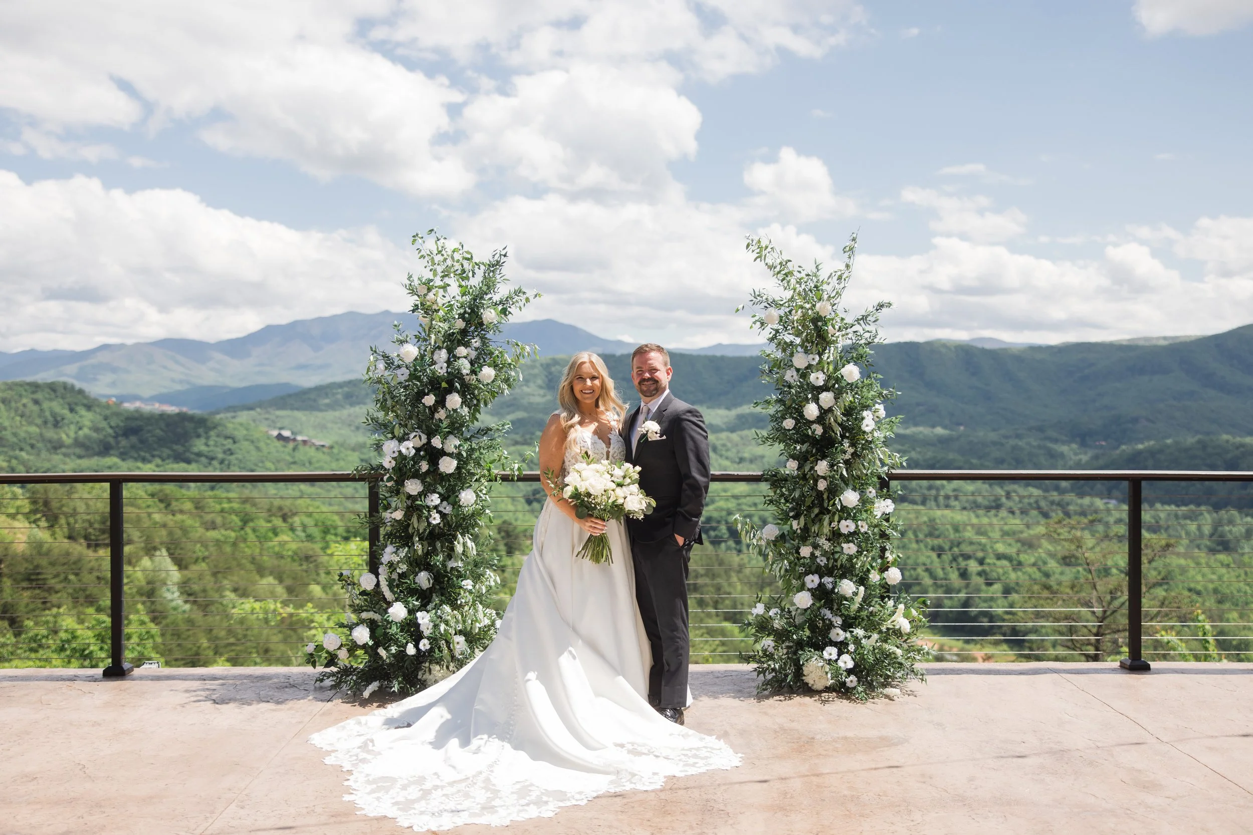 A bride and groom standing outdoors on a wedding day, surrounded by floral arrangements with white roses and greenery, with mountains and a cloudy sky in the background.