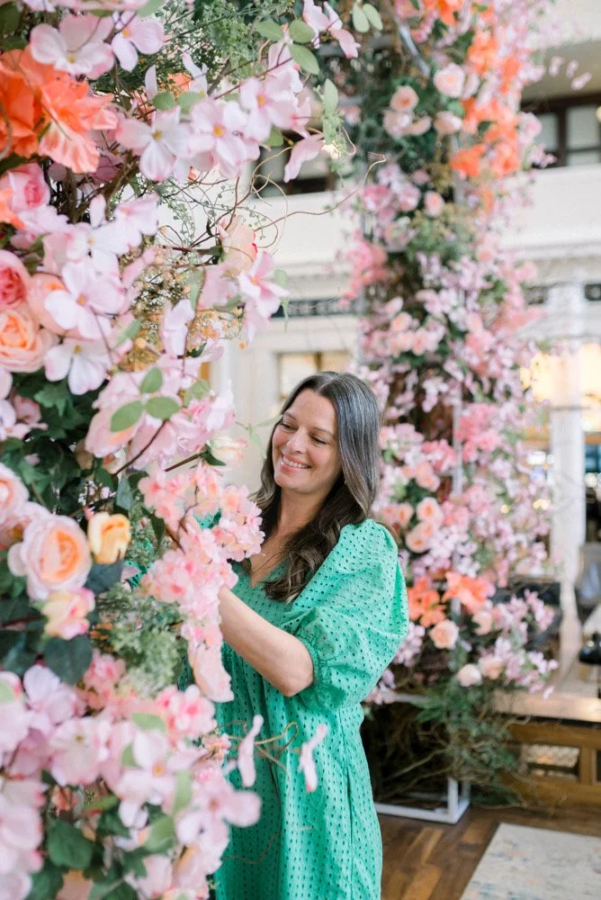 A woman in a green dress is standing amidst pink and peach flowers, smiling and arranging them in an indoor setting.