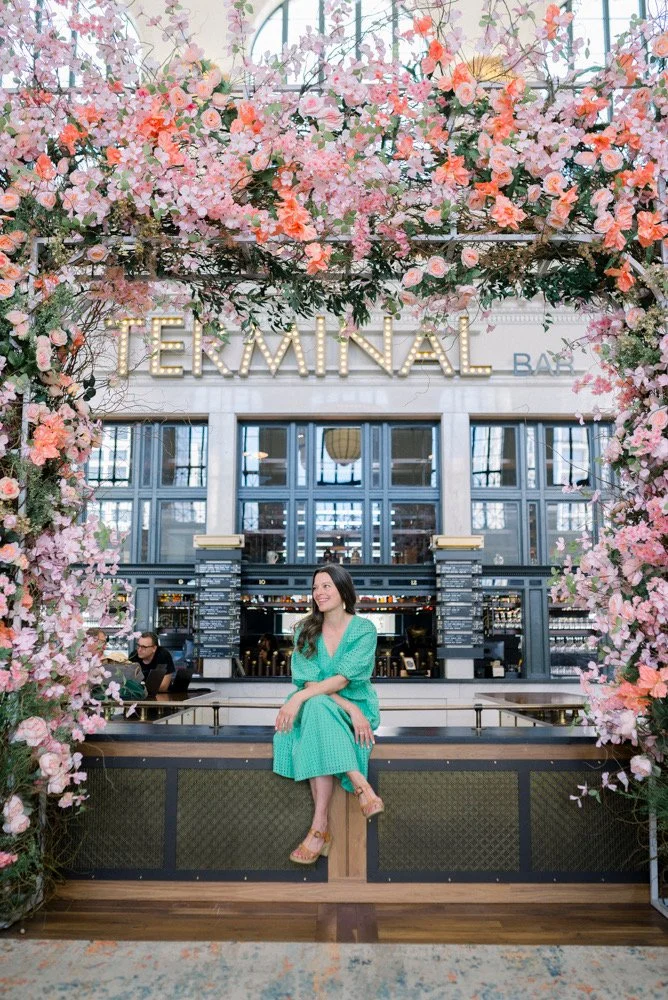 A woman in a turquoise dress sitting on a ledge in front of a floral arch inside a terminal building, with a sign above that reads 'TERMINAL'.