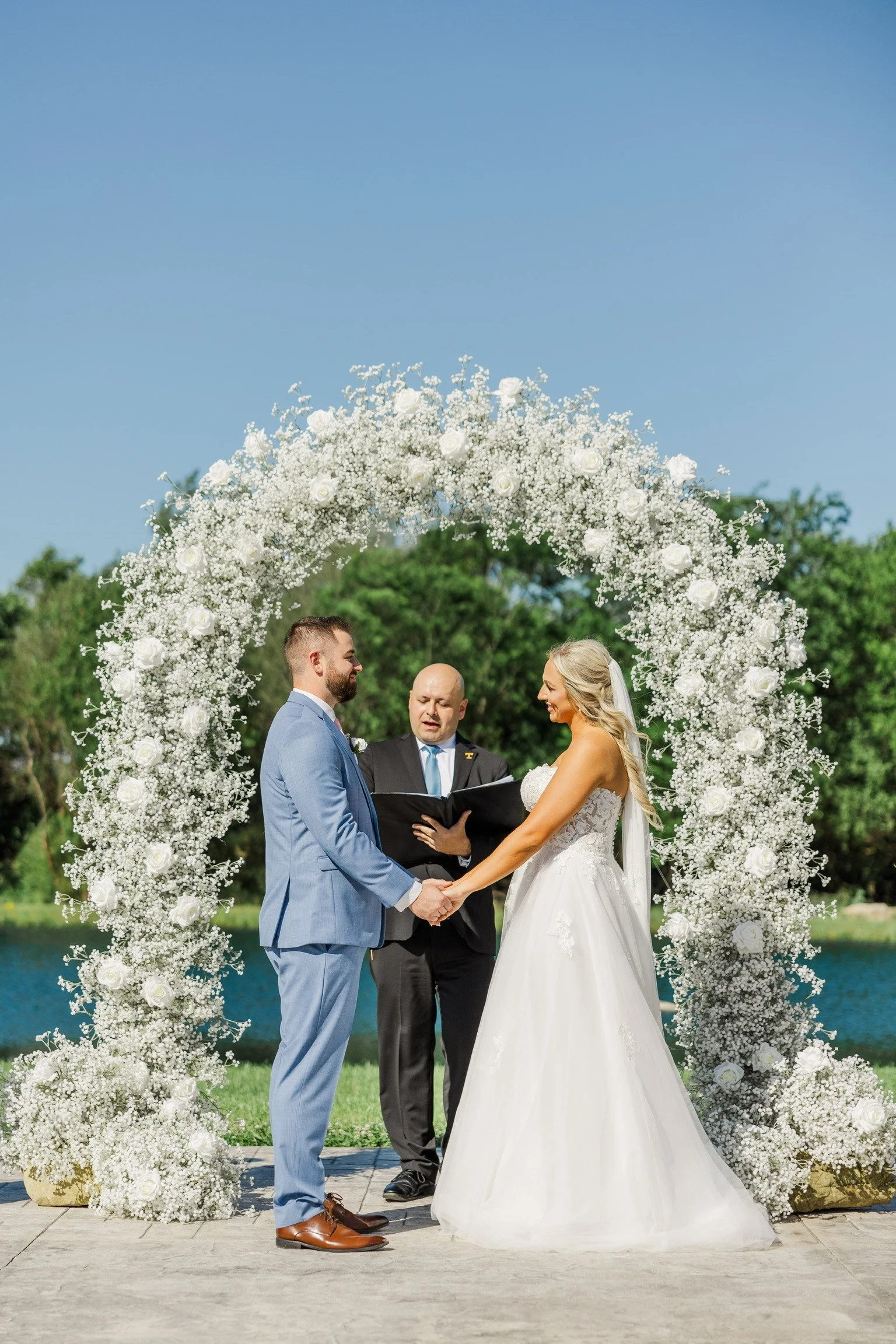 A bride and groom holding hands during their wedding ceremony outdoors, with an officiant reading, under a large arch made of white flowers with a lake and green trees in the background.