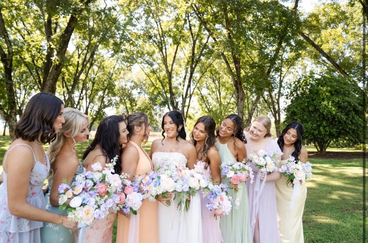 A bride and eight bridesmaids standing outdoors among trees, holding bouquets of flowers.