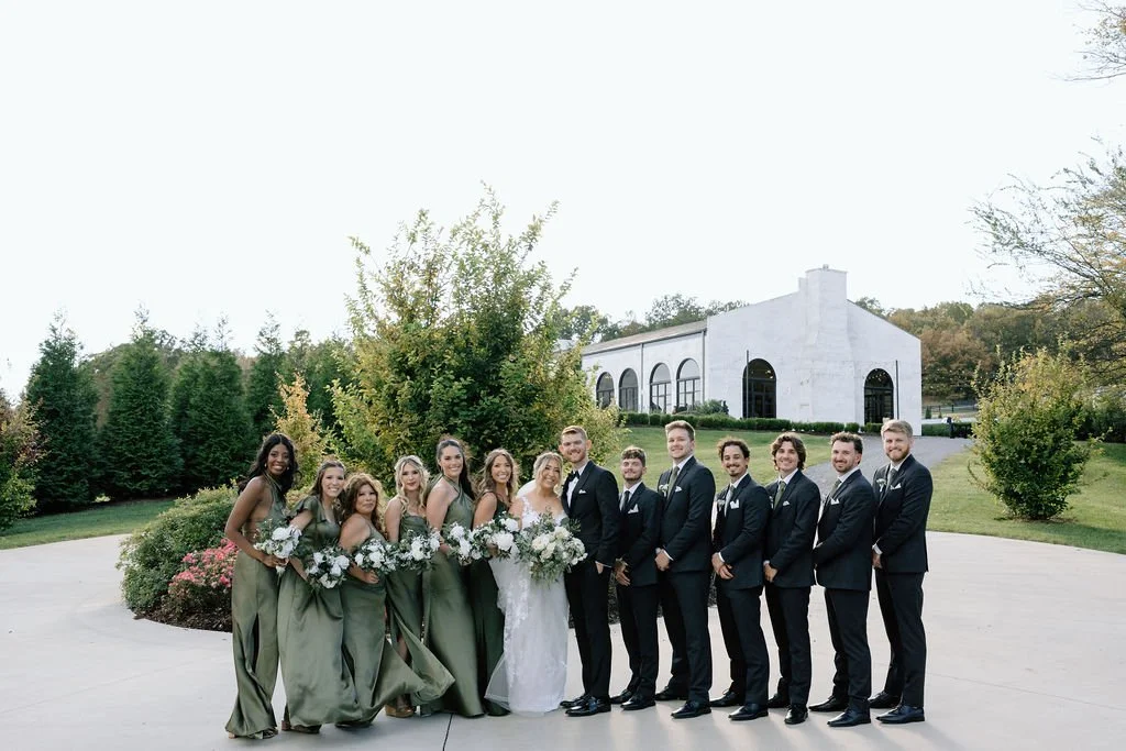 A wedding party standing outside in front of a white building, with the bride and groom in the center, surrounded by bridesmaids and groomsmen.