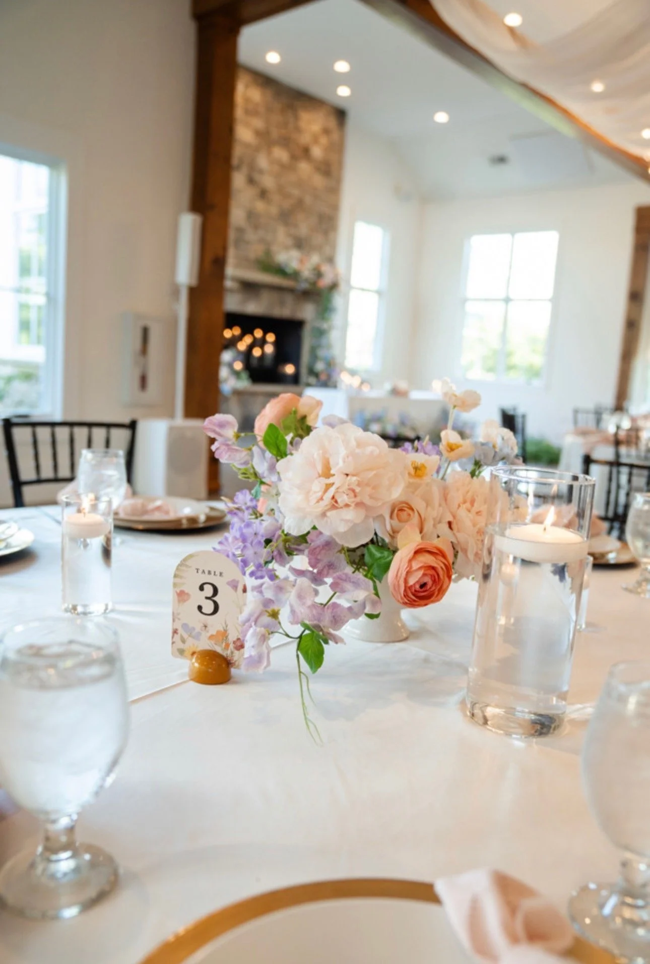 A close-up of a floral centerpiece on a dining table set for an event, with candles and a table number. The background shows a cozy room with large windows, a stone fireplace, and warm lighting.