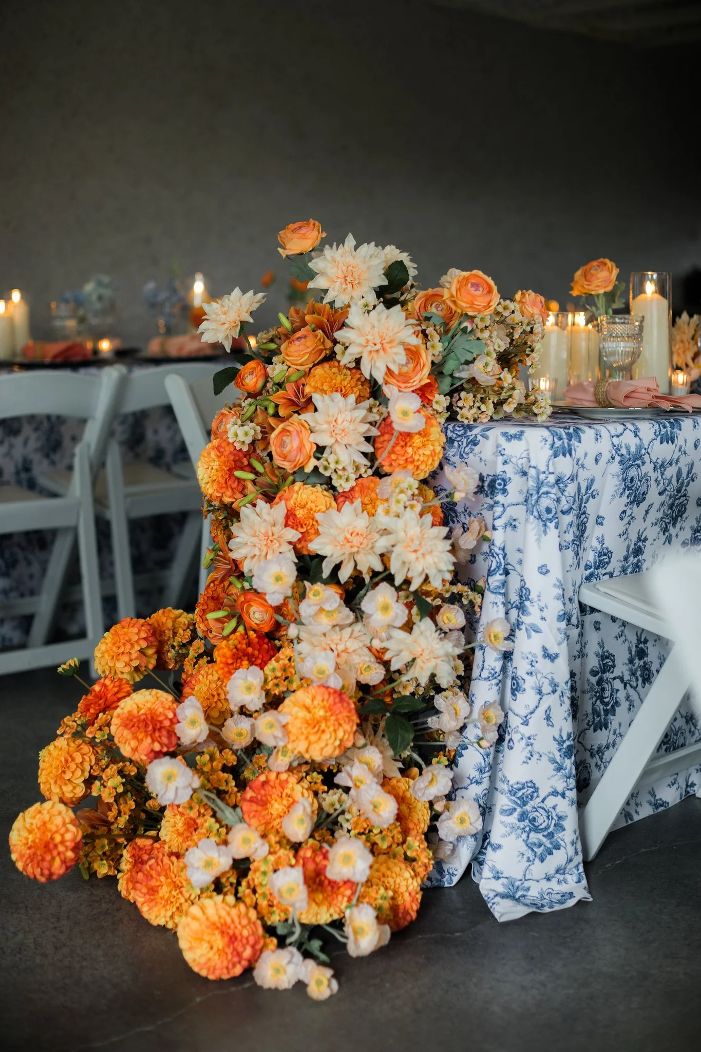 A floral centerpiece with orange and white flowers on a table covered with a blue and white patterned tablecloth, set for a special occasion with candles and pink napkins.