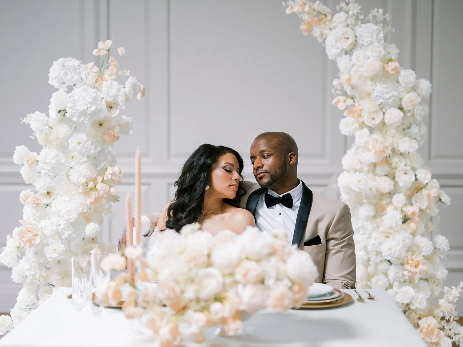 A newlywed couple sitting closely at their wedding reception table surrounded by an arch of white flowers and soft pastel blooms, with elegant table settings and a white paneled wall in the background.