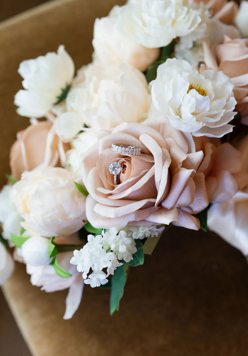 A close-up of a bouquet of white and blush flowers with a diamond engagement ring and wedding band resting on a blush rose.