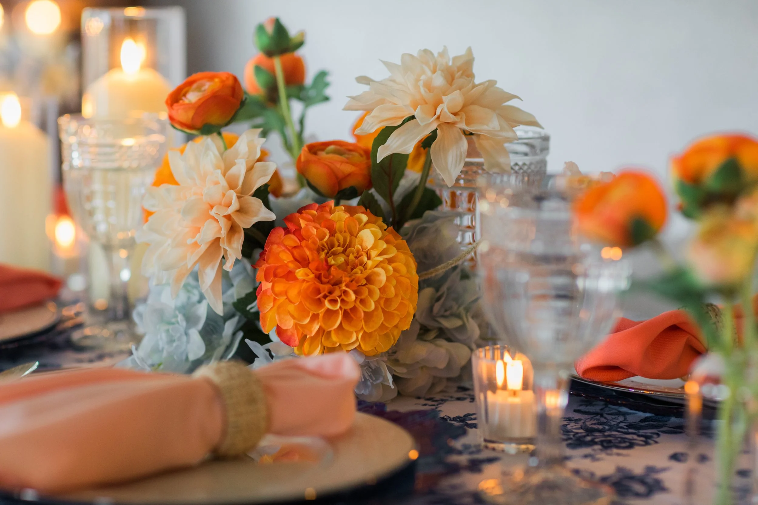 Close-up of a floral centerpiece with white, orange, and yellow flowers and lit candles in the background on a decorated table.