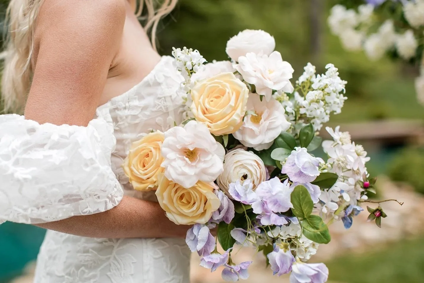 A woman in a white lace dress holding a bouquet of cream, white, and pastel purple flowers outdoors.