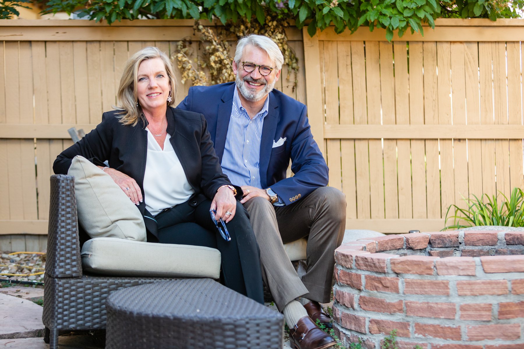 A smiling man and woman sitting together on outdoor furniture in a backyard, with a wooden fence and greenery in the background, near a brick fire pit.