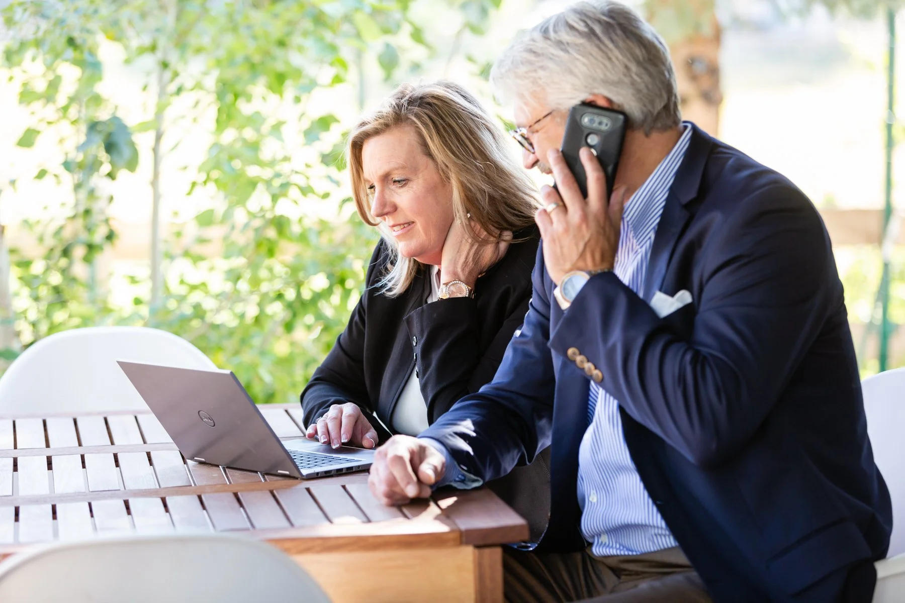 An older man in a navy suit talking on a mobile phone and a woman in a black blazer using a laptop at an outdoor table with greenery in the background.