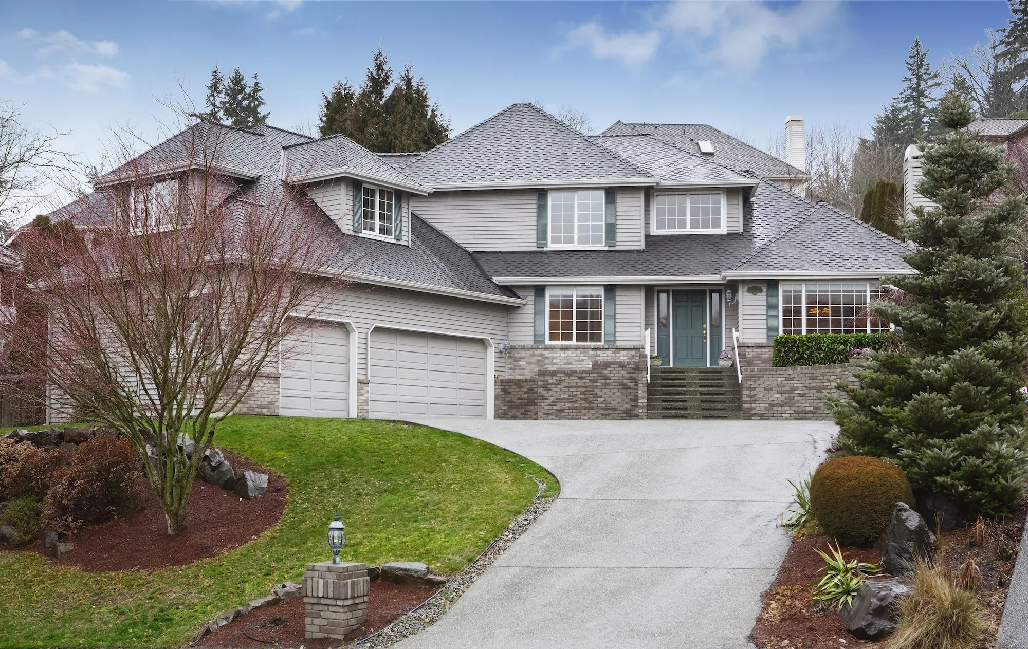 A large, two-story house with gray siding and a shingled roof, surrounded by landscaped yard with a curved walkway, trees, bushes, and decorative rocks.