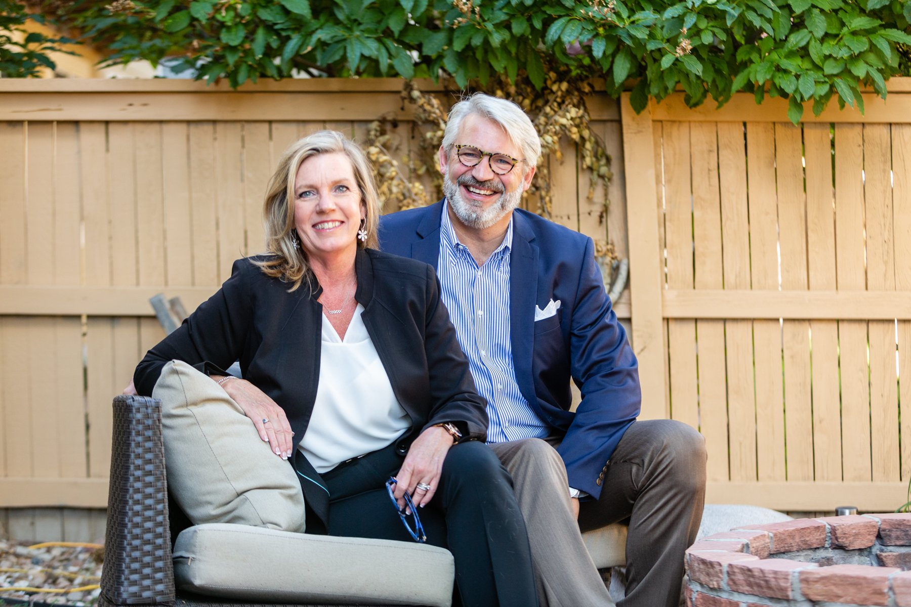 A smiling middle-aged couple sitting outside in a backyard with a wooden fence, green foliage, and a brick fire pit.