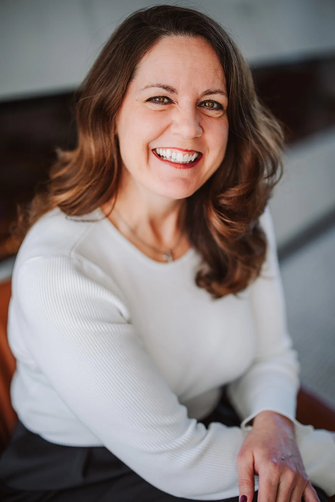 A smiling woman with shoulder-length brown hair, wearing a white long-sleeve shirt, seated and looking up at the camera.