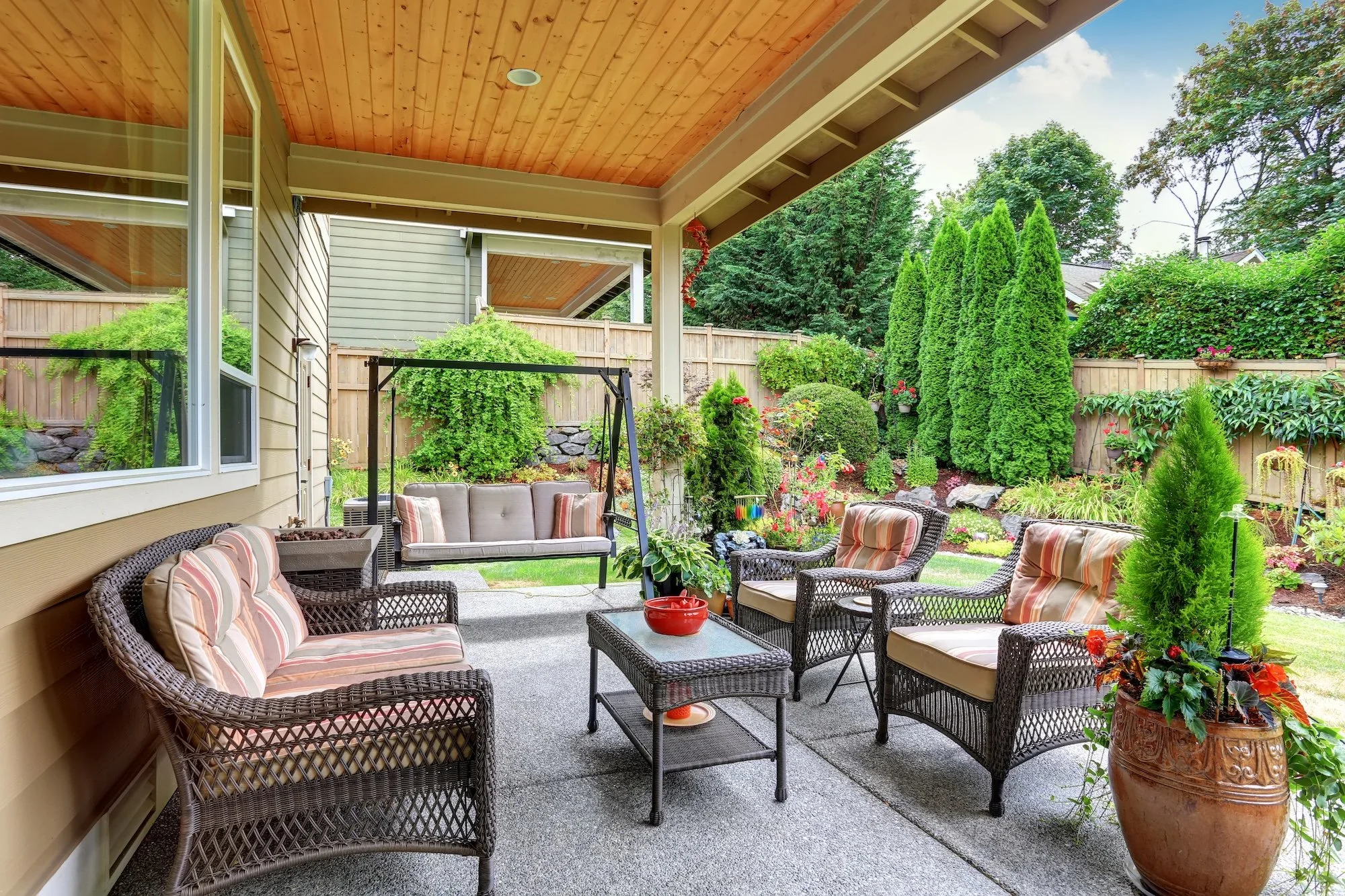 Patio with wicker furniture, cushions, potted plants, and lush green garden with trees and bushes in the background.